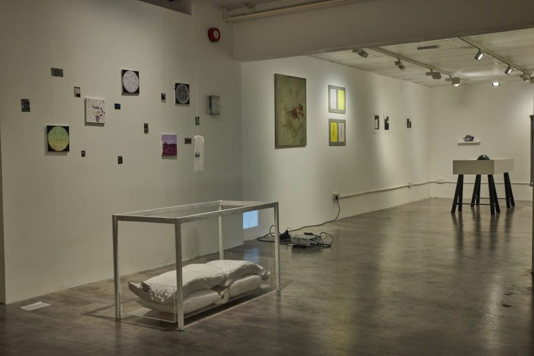 A contemporary gallery space with white walls displays small artworks arranged in a grid, along with larger framed pieces and a glass case containing two pillows. In the background, a white pedestal with a dark object on top stands near the far wall.