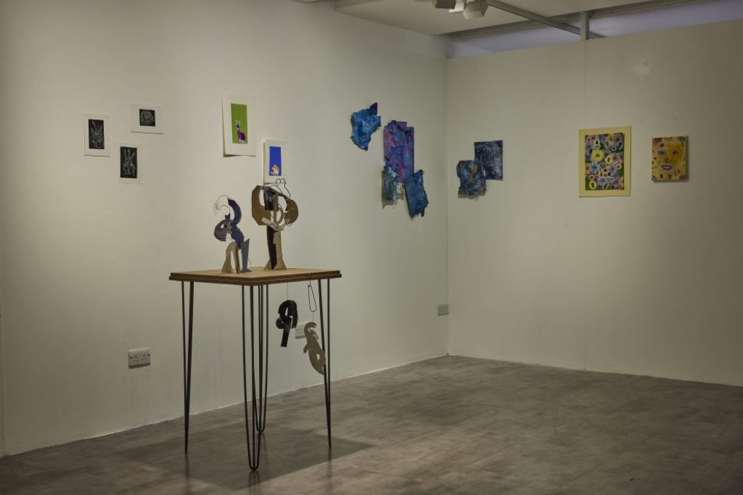 A gallery corner displays colorful abstract artworks on white walls, including small framed pieces and textured blue shapes. In the center, a wooden table with thin black legs holds sculptural objects made of cut-out materials.