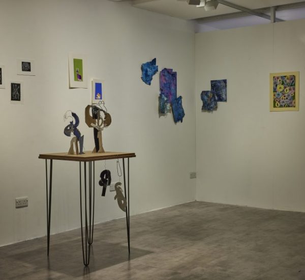 A gallery corner displays colorful abstract artworks on white walls, including small framed pieces and textured blue shapes. In the center, a wooden table with thin black legs holds sculptural objects made of cut-out materials.