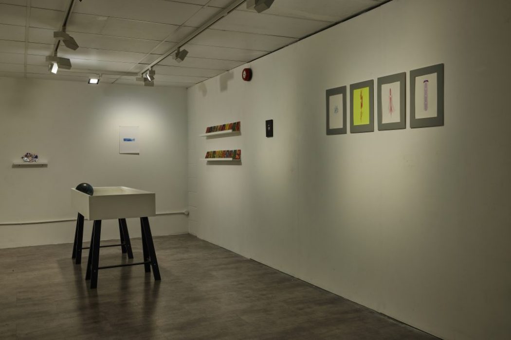 A minimalist gallery space with white walls displays small framed artworks and colorful pieces on shelves. A white pedestal with a black object on top stands in the center of the room under track lighting.