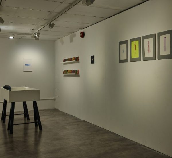 A minimalist gallery space with white walls displays small framed artworks and colorful pieces on shelves. A white pedestal with a black object on top stands in the center of the room under track lighting.