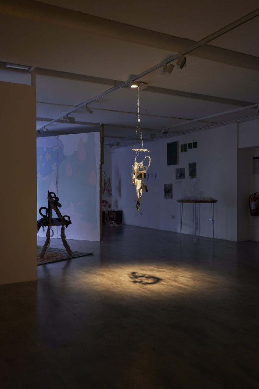 A dimly lit gallery space features a hanging sculpture illuminated by a spotlight, casting a shadow on the floor. In the background, additional artworks and installations are visible against white walls.