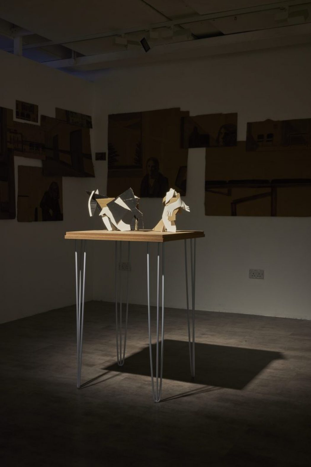 A wooden table with thin metal legs displays abstract sculptural pieces made of light-colored materials, illuminated by a spotlight. The background wall features several dark-toned artworks arranged in a collage-like manner.
