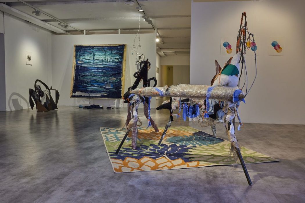 A gallery installation features an abstract sculptural piece made of mixed materials on a colorful floral-patterned rug in the foreground. In the background, a large blue textured artwork hangs on the wall alongside other abstract sculptures and small framed pieces.