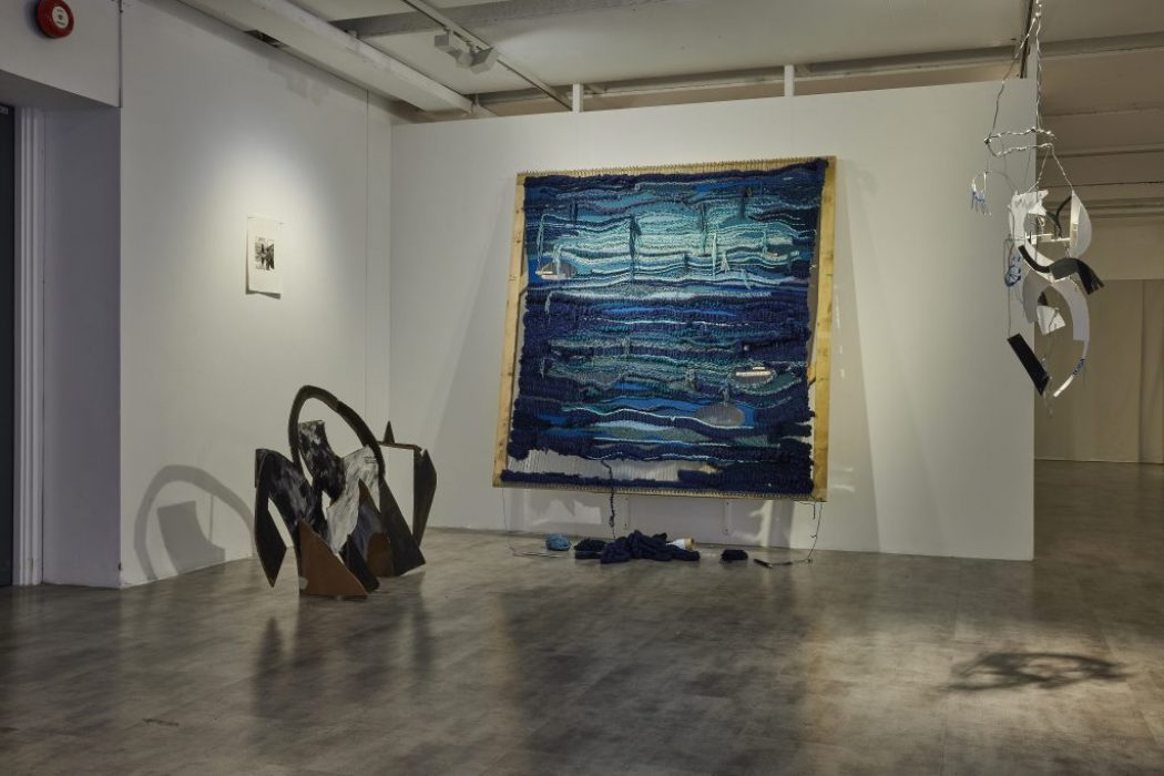 A gallery installation features a large textured artwork in shades of blue mounted on a white wall, with pieces of fabric on the floor beneath it. To the left is a dark abstract sculpture, and to the right, a hanging mobile made of curved metallic and black elements.