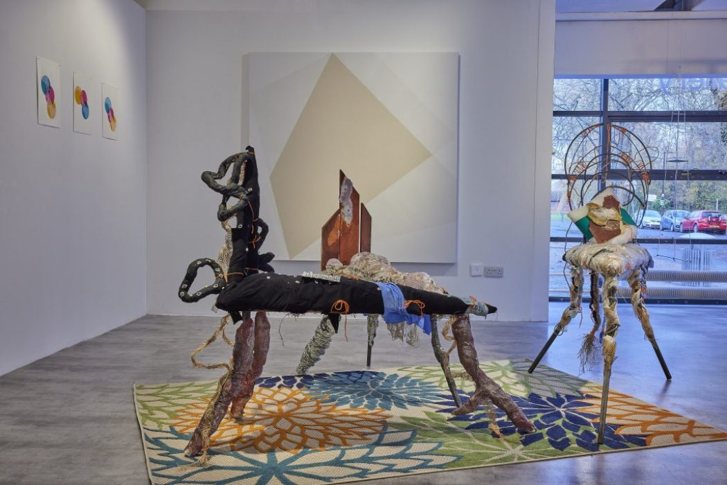 A gallery installation features two abstract sculptural pieces made of mixed materials positioned on a colorful floral-patterned rug. Behind them, a large geometric painting hangs on the wall, with three smaller circular artworks to the left and a window to the right.