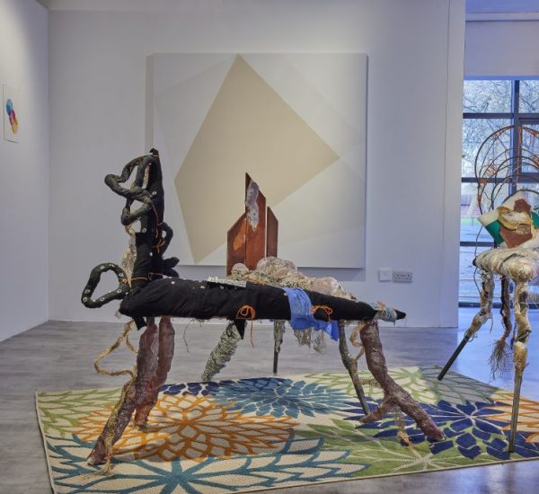 A gallery installation features two abstract sculptural pieces made of mixed materials positioned on a colorful floral-patterned rug. Behind them, a large geometric painting hangs on the wall, with three smaller circular artworks to the left and a window to the right.