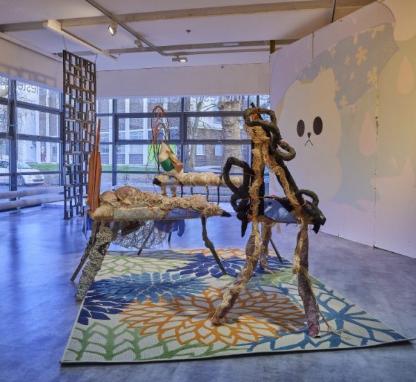 A contemporary gallery installation features abstract sculptural pieces made of textured materials arranged on a colorful floral-patterned rug. The background includes large windows and a wall mural with soft pastel shapes and a cartoon-like face.