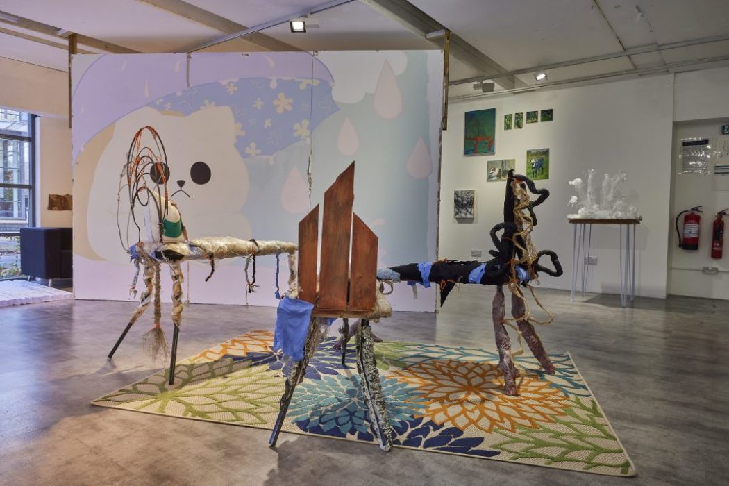 A gallery installation features two abstract sculptural pieces made of mixed materials placed on a colorful floral-patterned rug. Behind them, a large wall mural with a cartoon-like face and pastel shapes is visible, along with smaller artworks and a white sculptural piece on a pedestal.