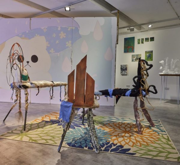 A gallery installation features two abstract sculptural pieces made of mixed materials placed on a colorful floral-patterned rug. Behind them, a large wall mural with a cartoon-like face and pastel shapes is visible, along with smaller artworks and a white sculptural piece on a pedestal.