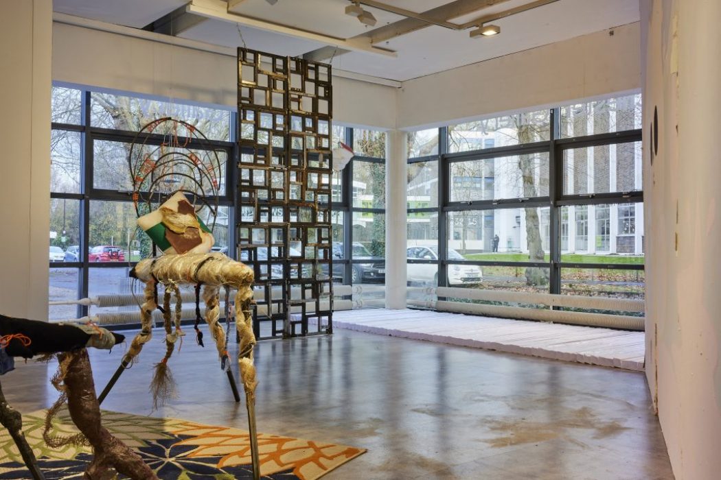 A gallery space with large windows features an abstract sculptural piece made of mixed materials positioned on a colorful patterned rug. In the background, a tall grid-like structure made of square frames stands near the windows, allowing natural light to fill the room.