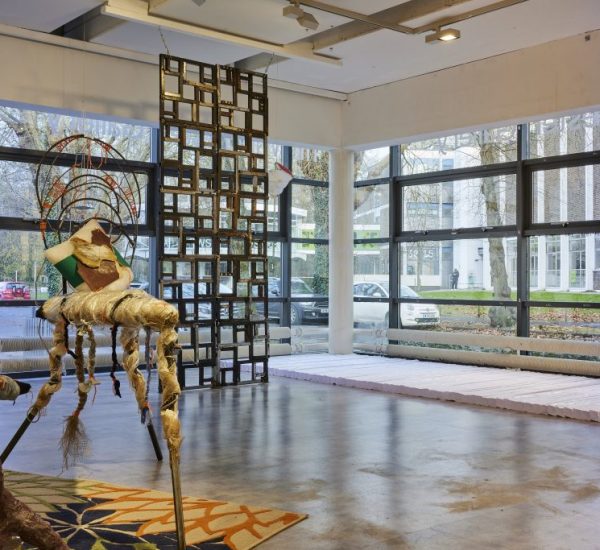 A gallery space with large windows features an abstract sculptural piece made of mixed materials positioned on a colorful patterned rug. In the background, a tall grid-like structure made of square frames stands near the windows, allowing natural light to fill the room.