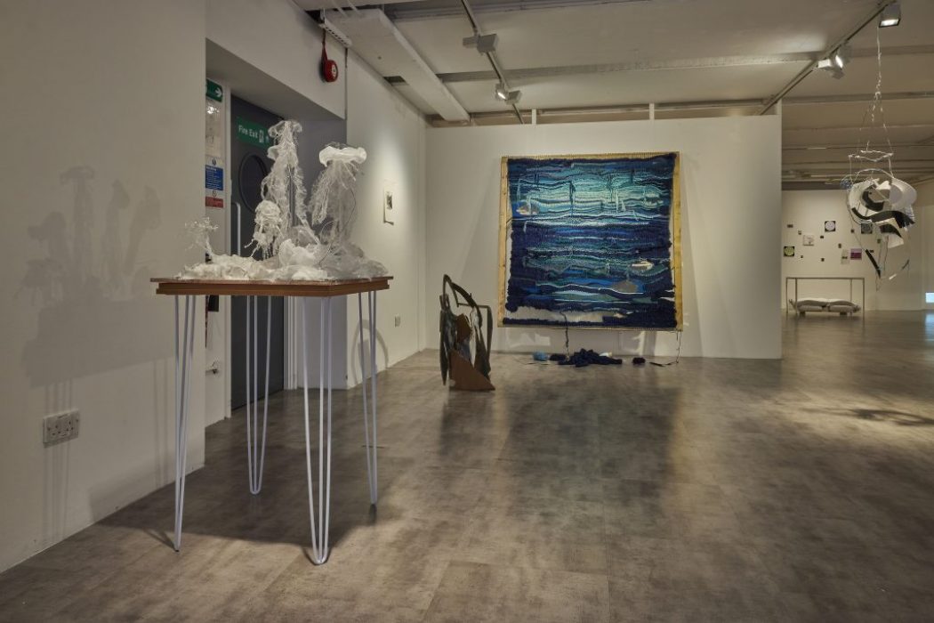 An art gallery with a white-walled interior displays various contemporary artworks, including a translucent sculpture on a tall table, a large blue textile piece on the back wall, and hanging abstract installations. The floor is gray and the space is lit with ceiling spotlights.