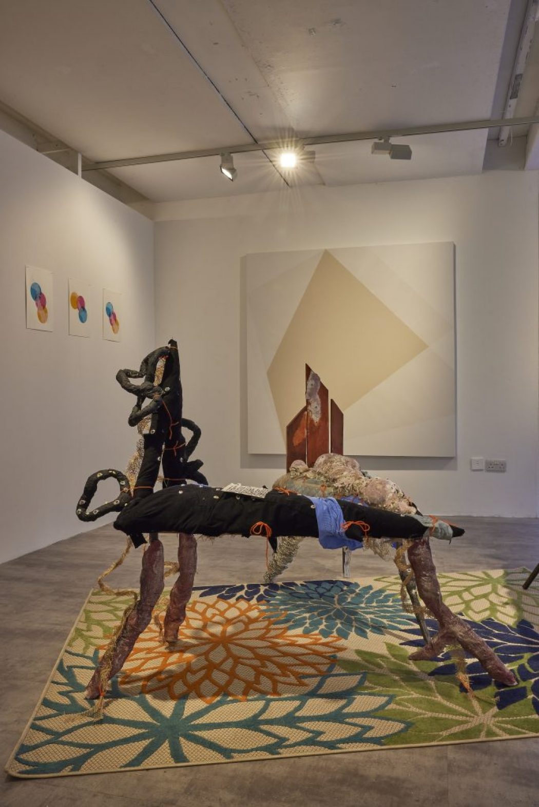 A gallery installation features two abstract sculptural pieces made of mixed materials positioned on a colorful floral-patterned rug. Behind them, a large geometric painting hangs on the wall, with three smaller circular artworks to the left and a window to the right.