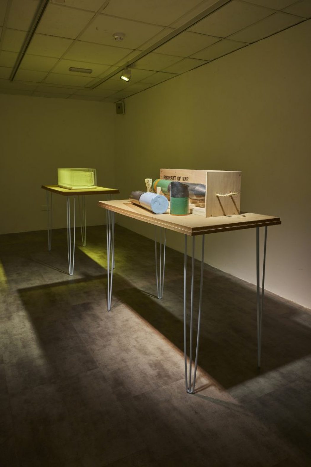 Two wooden tables with metal legs display small sculptural objects, including cylindrical shapes and a wooden box labeled “REMNANT OF WAR.” The room is dimly lit with a spotlight highlighting the tables against a plain wall.