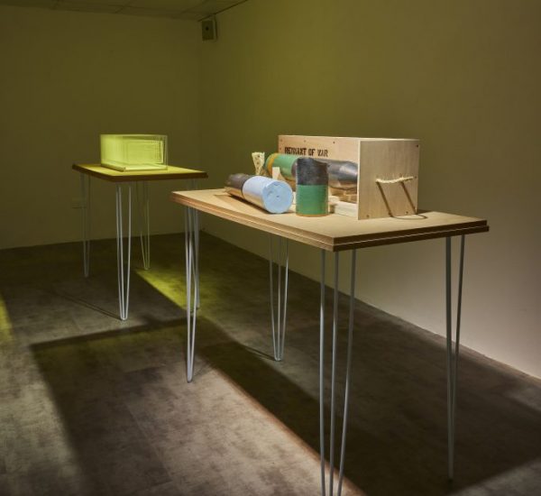 Two wooden tables with metal legs display small sculptural objects, including cylindrical shapes and a wooden box labeled “REMNANT OF WAR.” The room is dimly lit with a spotlight highlighting the tables against a plain wall.