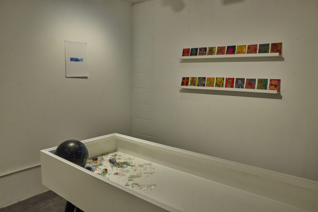 A white rectangular display box contains scattered colorful beads and a black sphere, with two rows of small, brightly colored artworks mounted on the wall behind it. A single blue-toned print hangs on the adjacent wall.