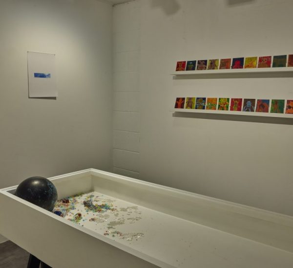 A white rectangular display box contains scattered colorful beads and a black sphere, with two rows of small, brightly colored artworks mounted on the wall behind it. A single blue-toned print hangs on the adjacent wall.