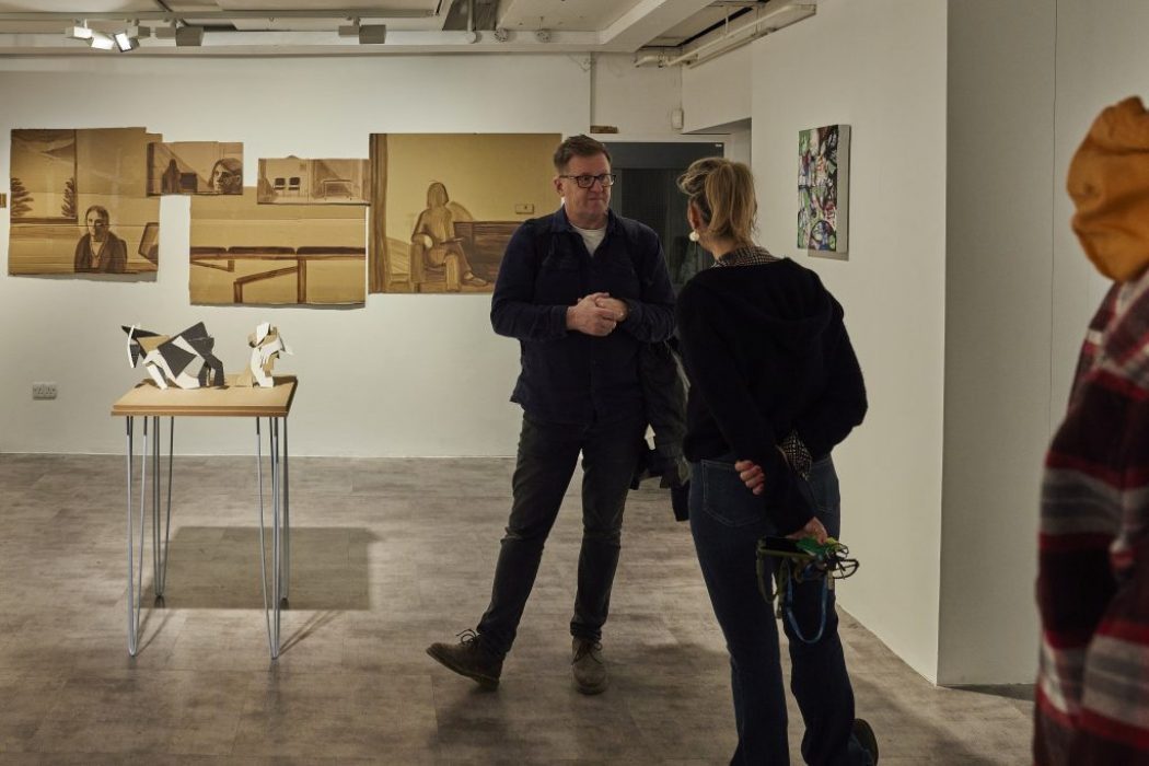 Two people stand in an art gallery near a wooden table displaying abstract black and white sculptures, with several sepia-toned portraits and interior scenes mounted on the wall behind them. Another colorful artwork is visible on the adjacent wall, and part of a third person is seen on the right edge of the image.