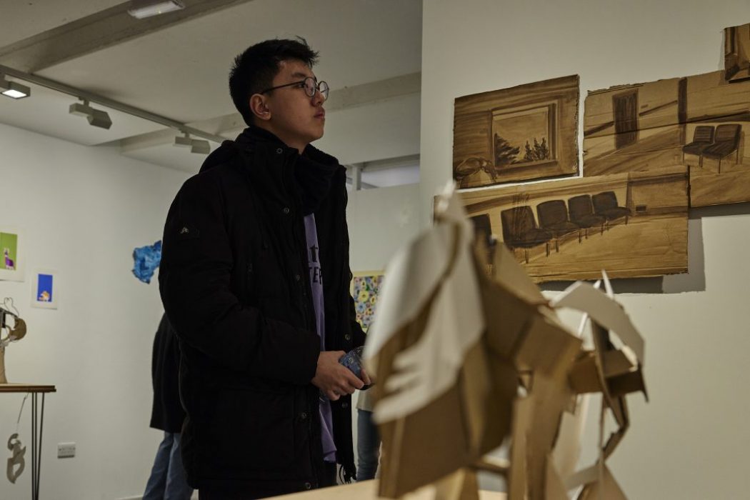 A person in a dark jacket stands in an art gallery, looking at brown-toned artworks on the wall depicting interior scenes with chairs. In the foreground, there is a sculptural piece made of layered cardboard shapes.
