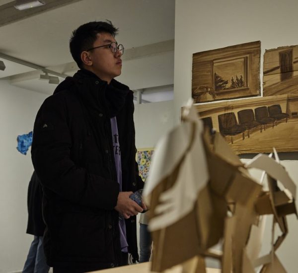 A person in a dark jacket stands in an art gallery, looking at brown-toned artworks on the wall depicting interior scenes with chairs. In the foreground, there is a sculptural piece made of layered cardboard shapes.