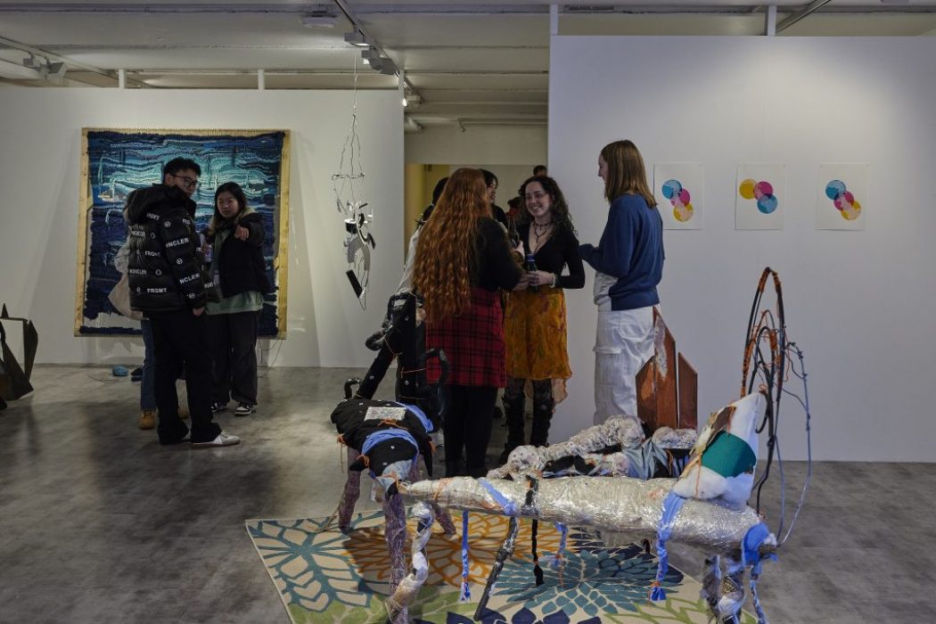 A group of people stand and talk in a contemporary art gallery featuring colorful abstract artworks on the walls and mixed-media sculptures on the floor. The space includes a large blue textile piece, hanging installation, and a patterned rug with sculptural elements.