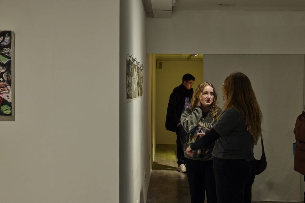 Two people stand in a narrow gallery hallway looking at small artworks displayed on the wall, while another person is visible further back in the corridor. A single framed piece hangs on the left wall, and the space is lit with overhead lighting.