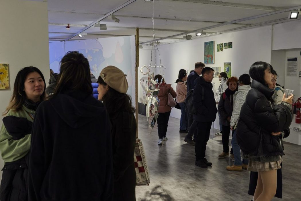 A group of people are gathered in a contemporary art gallery, viewing and discussing artworks displayed on white walls. The space features hanging sculptures, framed pieces, and colorful paintings arranged throughout the room.