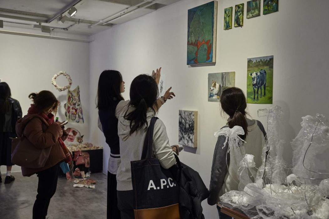Several people are viewing and discussing artworks displayed on a white gallery wall, which includes paintings of landscapes, animals, and abstract designs. Sculptural pieces made of translucent material and other mixed-media works are arranged on tables and the floor nearby.