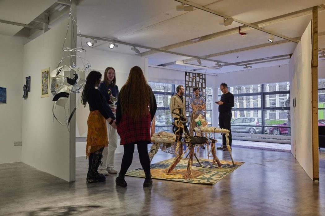 A group of people stand and talk in a contemporary art gallery featuring mixed-media sculptures arranged on a patterned rug in the center. The space includes hanging installations, framed artworks on white walls, and large windows in the background letting in natural light.