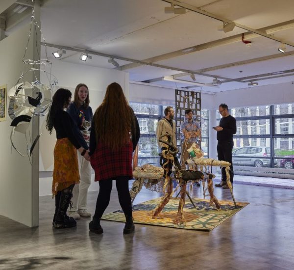A group of people stand and talk in a contemporary art gallery featuring mixed-media sculptures arranged on a patterned rug in the center. The space includes hanging installations, framed artworks on white walls, and large windows in the background letting in natural light.