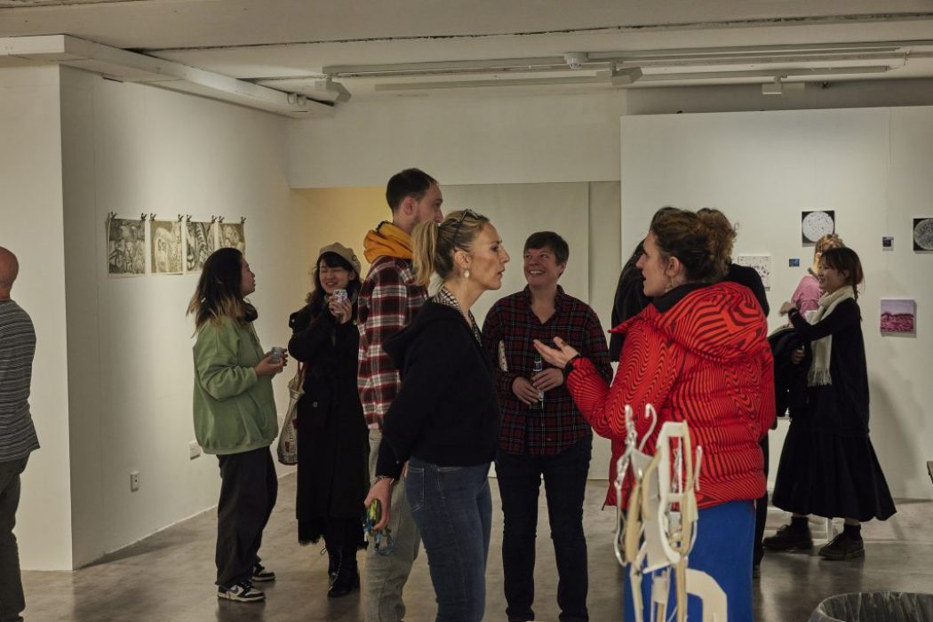 A group of people stand and talk in a contemporary art gallery with white walls displaying small framed artworks and drawings. The space is well-lit with track lighting, and some sculptural elements are visible in the foreground.