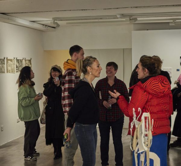 A group of people stand and talk in a contemporary art gallery with white walls displaying small framed artworks and drawings. The space is well-lit with track lighting, and some sculptural elements are visible in the foreground.