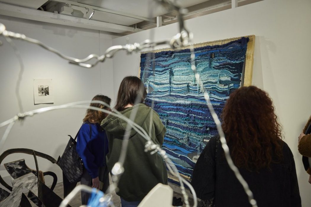 Three people stand closely viewing a large blue and turquoise woven textile artwork displayed on a white wall. The scene is partially framed by out-of-focus wire elements in the foreground, adding depth to the image.