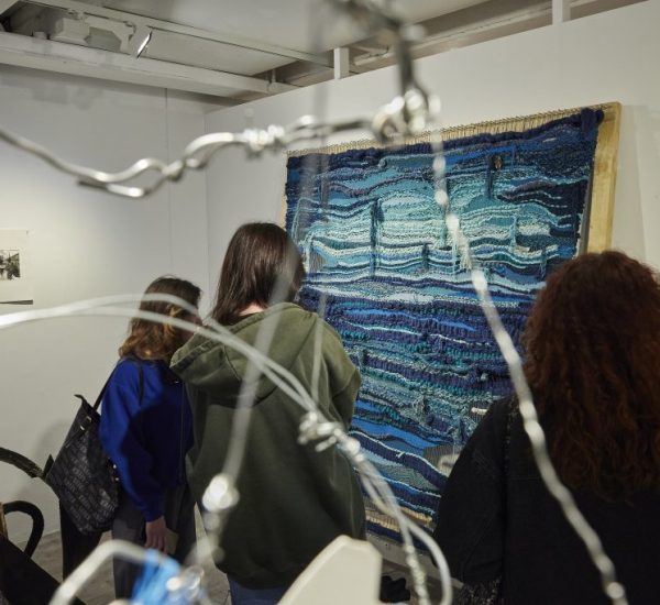 Three people stand closely viewing a large blue and turquoise woven textile artwork displayed on a white wall. The scene is partially framed by out-of-focus wire elements in the foreground, adding depth to the image.