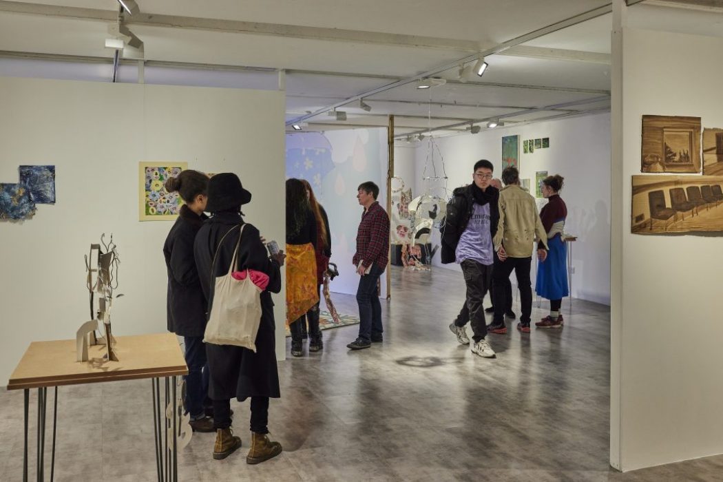 A group of people are gathered in a contemporary art gallery, viewing colorful paintings on the walls and sculptural pieces on display. The space features white walls, bright lighting, and a mix of hanging and freestanding artworks.