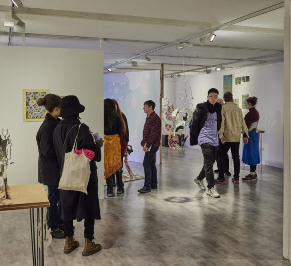 A group of people are gathered in a contemporary art gallery, viewing colorful paintings on the walls and sculptural pieces on display. The space features white walls, bright lighting, and a mix of hanging and freestanding artworks.