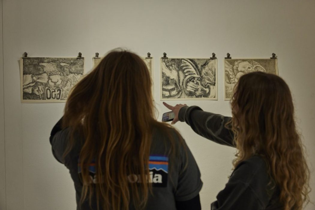 Two people stand in front of a white wall examining four monochrome drawings clipped in a row. One person is pointing at a drawing while holding a phone, and the artworks feature abstract and organic shapes.