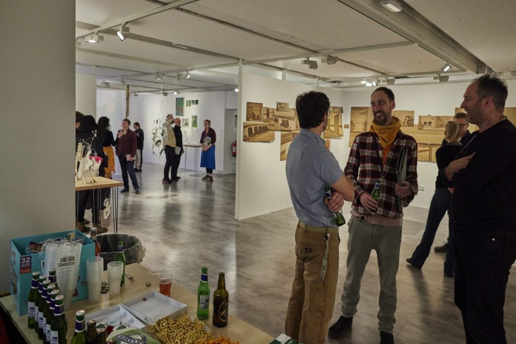 A gallery space with white walls displays architectural drawings and models, while several people stand and converse in small groups. In the foreground, a table holds drinks, snacks, and empty cups, suggesting a social event or exhibition opening.