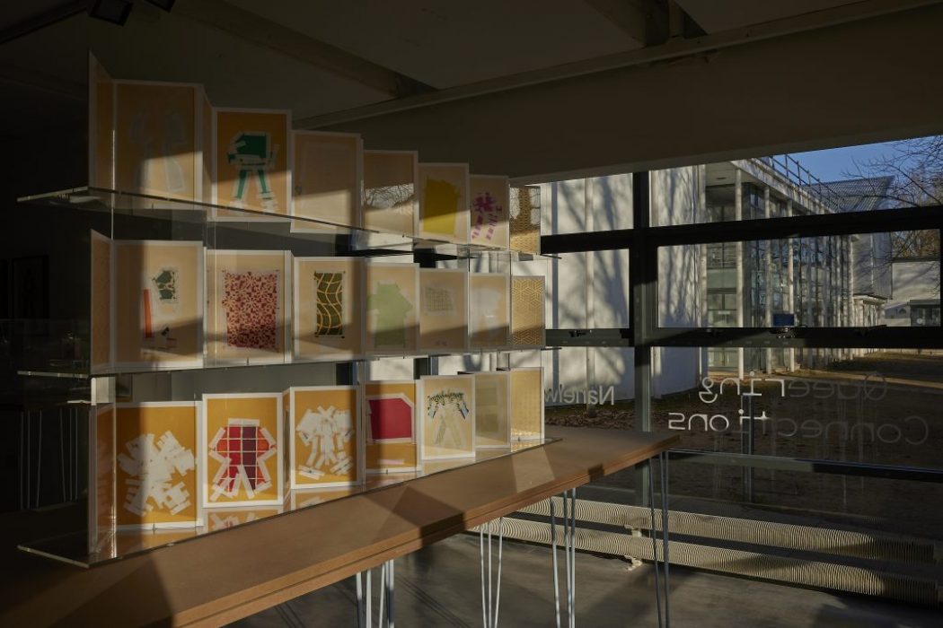 A display of colorful, layered paper artworks is arranged on clear shelves inside a gallery space, with sunlight streaming through large windows. The pieces feature abstract shapes and patterns in shades of yellow, orange, green, and red.