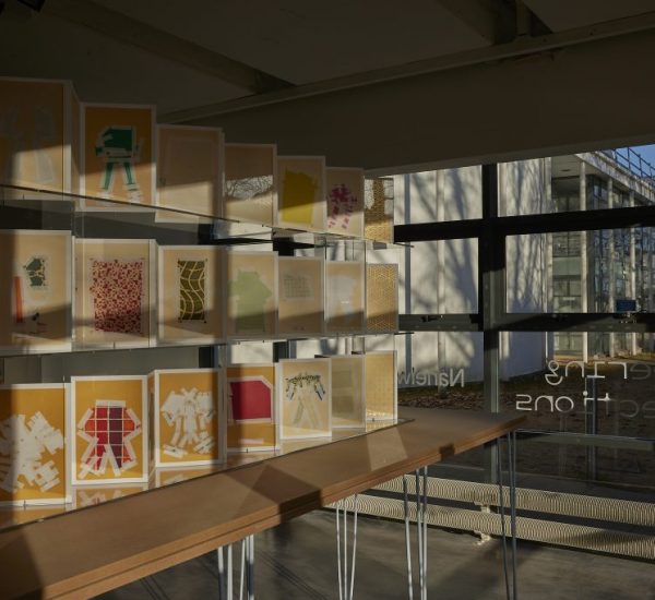 A display of colorful, layered paper artworks is arranged on clear shelves inside a gallery space, with sunlight streaming through large windows. The pieces feature abstract shapes and patterns in shades of yellow, orange, green, and red.