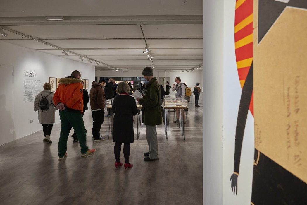 A group of people are gathered in a modern art gallery with white walls and track lighting. They are observing artworks displayed on both the walls and tables.