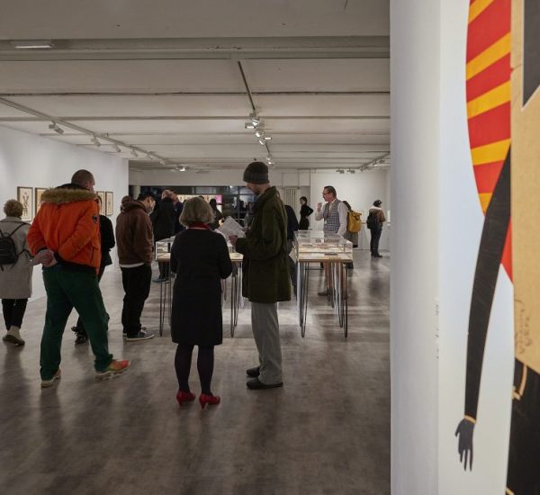 A group of people are gathered in a modern art gallery with white walls and track lighting. They are observing artworks displayed on both the walls and tables.
