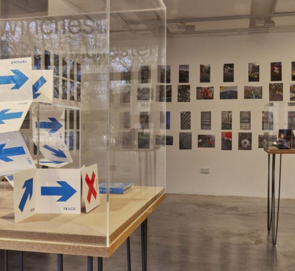An art gallery display features a folded paper structure marked with blue arrows and a red 'X' inside a glass case. Behind it, a wall is covered with small photographs arranged in a grid, and another table with items is visible to the right.