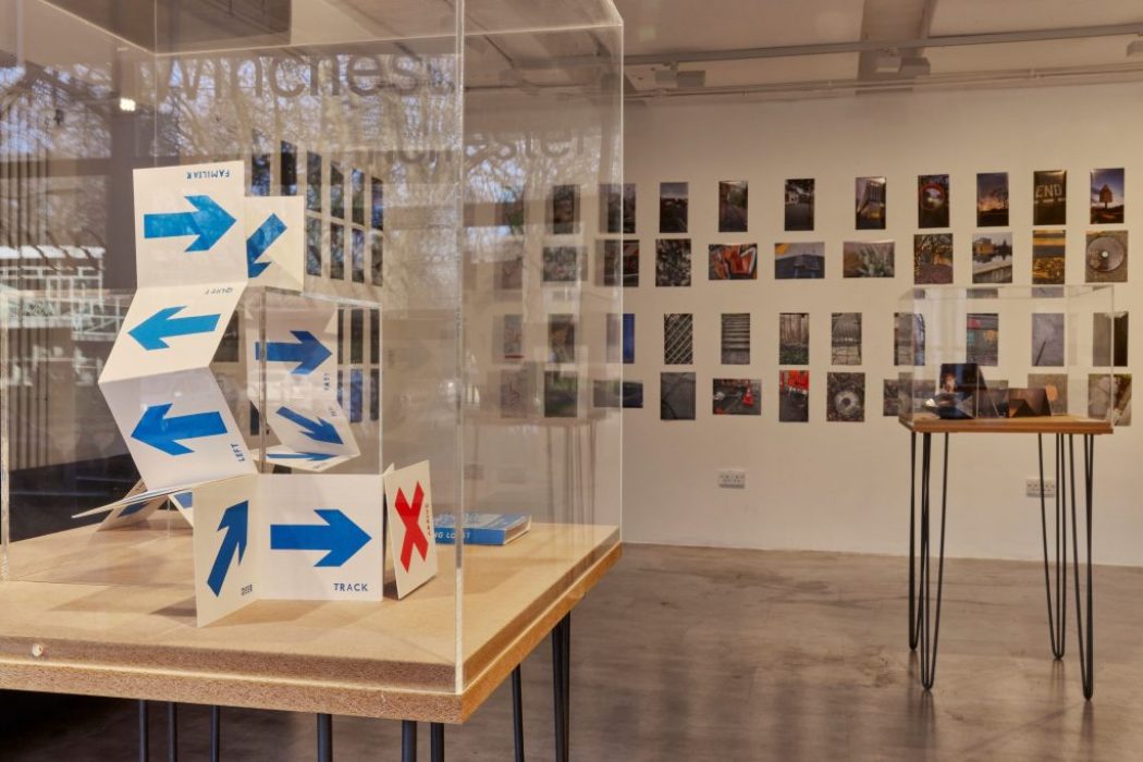 An art gallery display features a folded paper structure marked with blue arrows and a red 'X' inside a glass case. Behind it, a wall is covered with small photographs arranged in a grid, and another table with items is visible to the right.