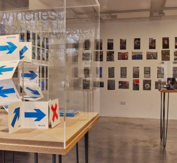 An art gallery display features a folded paper structure marked with blue arrows and a red 'X' inside a glass case. Behind it, a wall is covered with small photographs arranged in a grid, and another table with items is visible to the right.