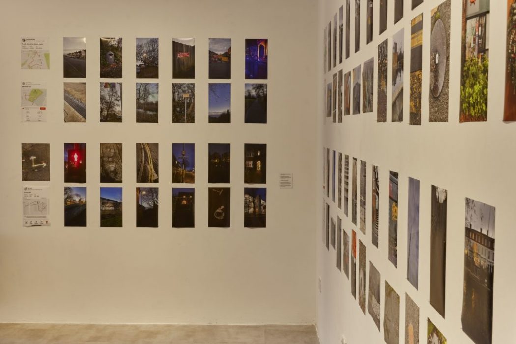 A gallery exhibition features numerous photographs arranged in a grid on two adjacent walls. The images depict a variety of scenes including landscapes, urban settings, and night views.
