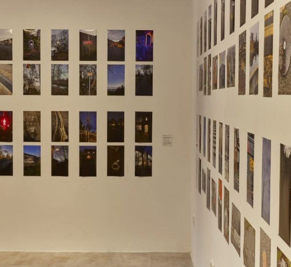 A gallery exhibition features numerous photographs arranged in a grid on two adjacent walls. The images depict a variety of scenes including landscapes, urban settings, and night views.