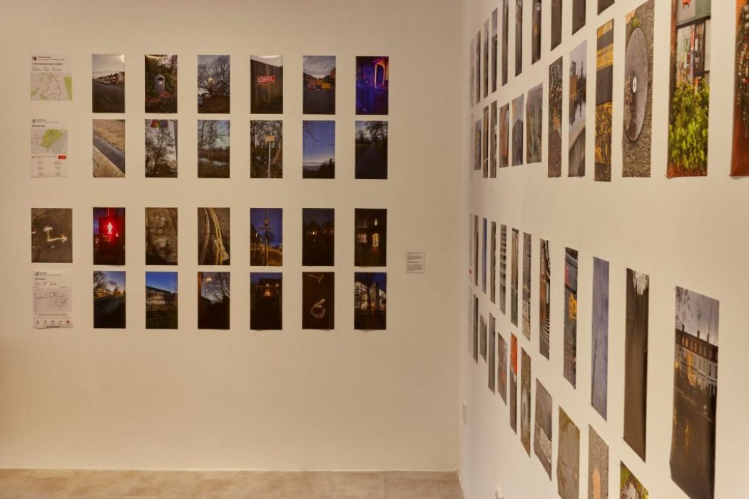 A gallery exhibition features numerous photographs arranged in a grid on two adjacent walls. The images depict a variety of scenes including landscapes, urban settings, and night views.