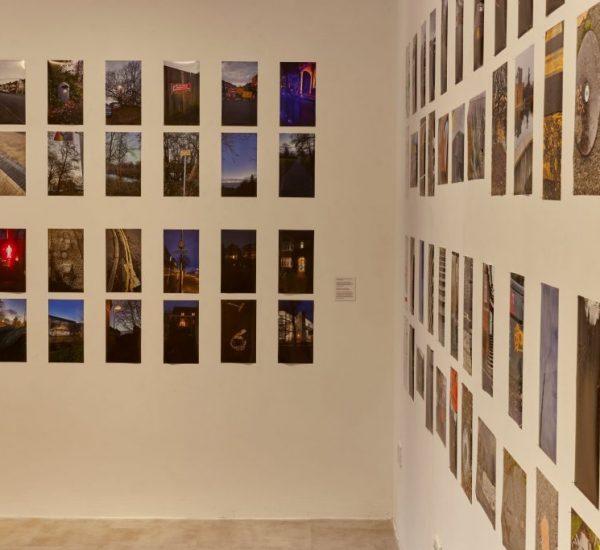 A gallery exhibition features numerous photographs arranged in a grid on two adjacent walls. The images depict a variety of scenes including landscapes, urban settings, and night views.